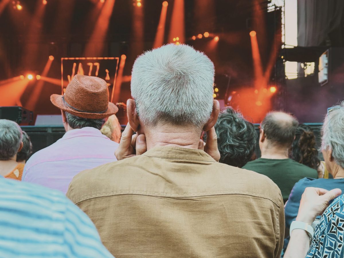 An old man plugging his ears with his fingers at a concert, seen from the back. Image Anthony Nelzin-Santos.